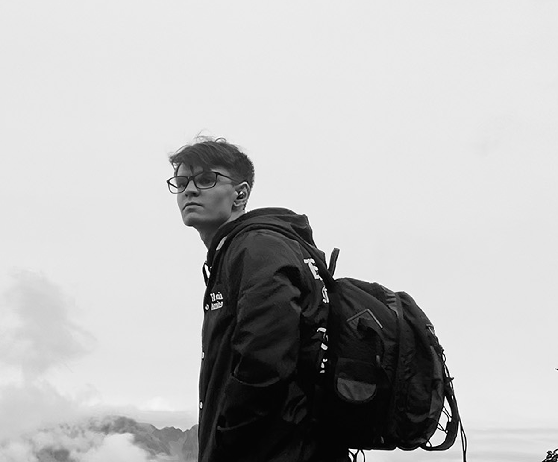 Portrait of Josh Sinkinson wearing glasses and a backpack, photographed on a mountain with misty clouds below